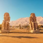 The two seated Colossi of Memnon in warm sunlight with palm trees and the west bank mountains behind