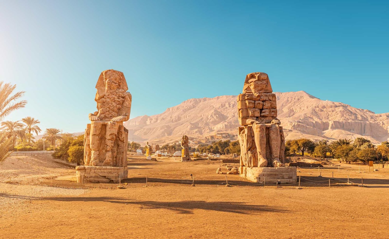 The two seated Colossi of Memnon in warm sunlight with palm trees and the west bank mountains behind